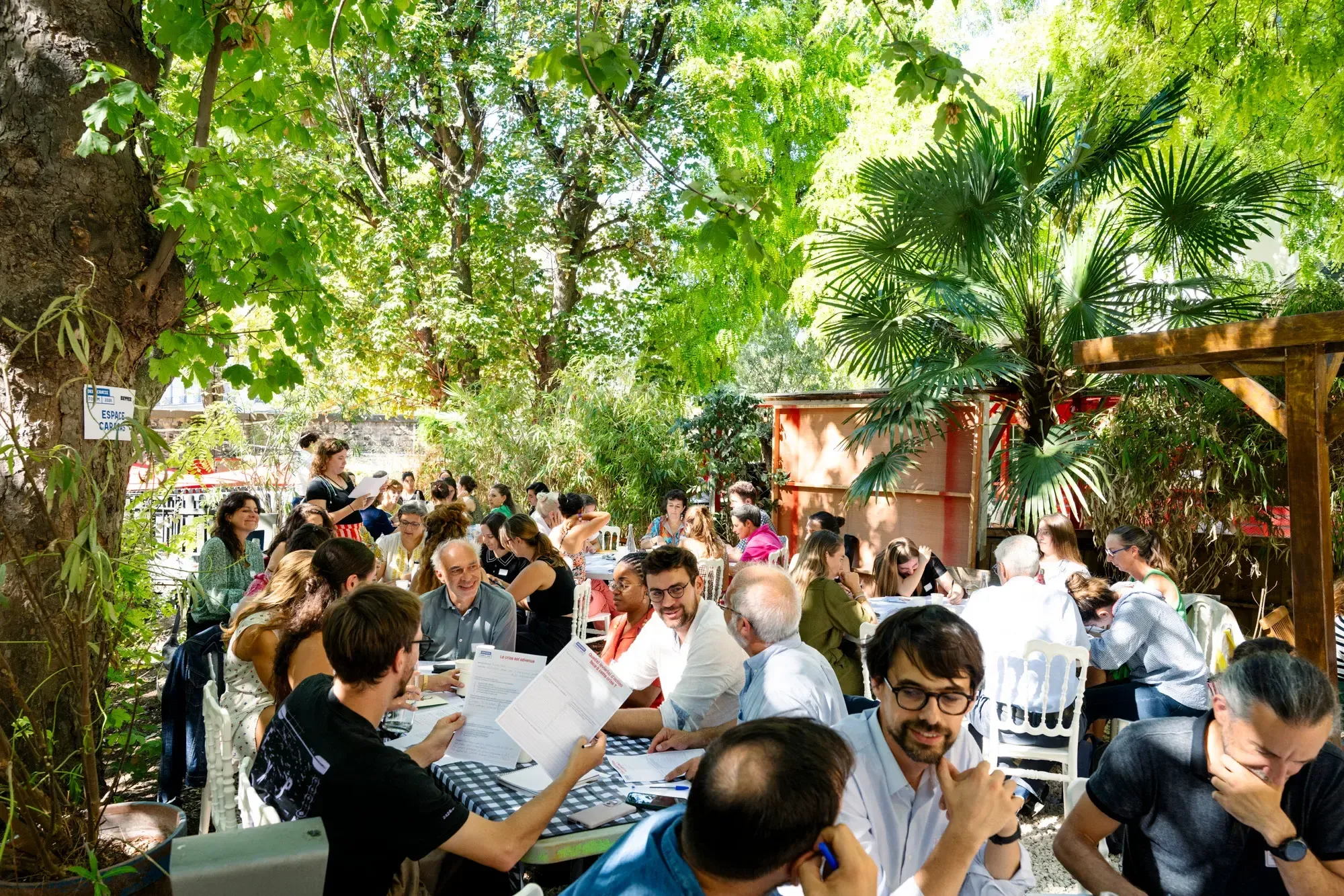 Des dizaines de personnes attablées sous les arbres du jardin, en atelier, près du panneau « Espace Caracas ».
